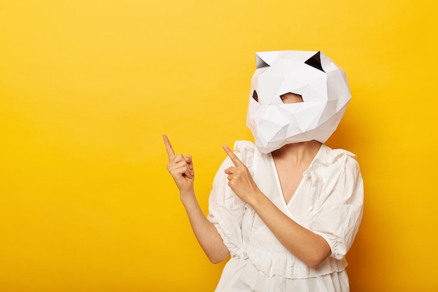 Innenaufnahme einer unbekannten Frau mit Kleid und Papierkatzenmaske, die auf den Werbebereich zeigt, Kopienraum für Werbung, die isoliert vor gelbem Hintergrund posiert