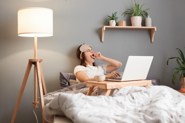 Innenaufnahme einer verschlafenen gelangweilten Frau mit Schlafmaske, die von zu Hause aus am Laptop arbeitet, während sie auf dem Bett liegt und lange Stunden vor dem Monitor verbringt, fühlt sich schläfrig gähnend und bedeckt den Mund mit der Handfläche