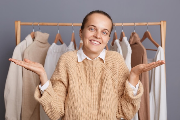 Innenaufnahme einer verwirrten, ahnungslosen Modestylistin, die Zweifel gestikuliert, welche Kleidung sie wählen soll, wenn sie in der Nähe eines Kleiderschranks mit trendigem Outfit auf einem Regal in einer Boutique steht