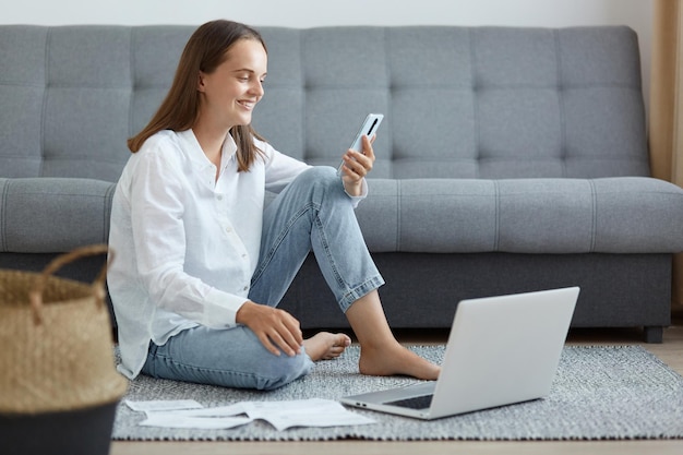 Innenaufnahme einer zufriedenen, erfreut lächelnden Frau mit weißem Hemd und Jeans, die auf dem Boden sitzt und ein Handy hält und mit einem charmanten Lächeln auf das Telefon schaut