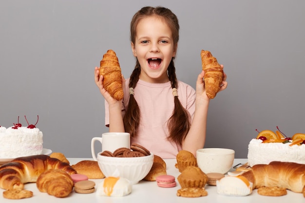 Innenaufnahme eines aufgeregten, erstaunten kleinen Mädchens mit Zöpfen, das isoliert vor grauem Hintergrund am Tisch sitzt und vor Glück und Aufregung mit Croissant schreit