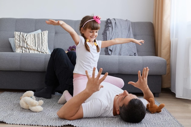 Innenaufnahme eines aufgeregten kleinen Mädchens und eines jungen erwachsenen Vaters, der auf dem Boden in der Nähe des Sofas in Freizeitkleidung spielt, die Familie aktiv Zeit miteinander verbringt und glücklich lacht.