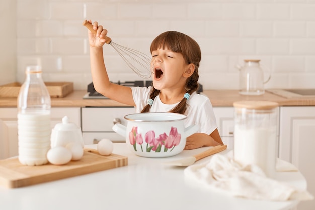 Innenaufnahme eines aufgeregten kleinen süßen weiblichen Kindes mit dunklem Haar, das ein weißes T-Shirt trägt, das in der hellen Küche kocht, die Krone in den Händen hält und beim Kochen wie ein Mikrofon singt.