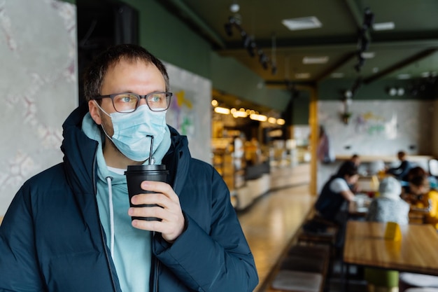 Innenaufnahme eines ernsten, nachdenklichen Mannes, der Kaffee zum Mitnehmen trinkt, trägt eine Schutzmaske, eine transparente Brillenjacke, die in einem überfüllten Café posiert, folgt den Quarantäneregeln während des Ausbruchs des Coronavirus