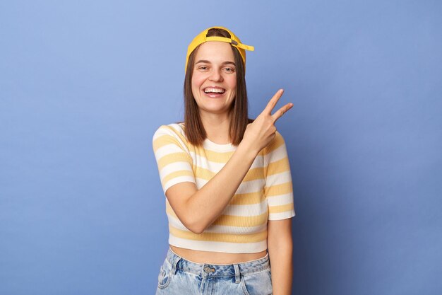 Innenaufnahme eines fröhlichen, fröhlichen Teenager-Mädchens mit gestreiftem T-Shirt und Baseballkappe, das isoliert vor blauem Hintergrund steht und die Siegesgeste des Vi-Zeichens zeigt, die mit einem zahnigen Lächeln in die Kamera blickt