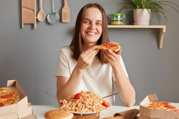 Innenaufnahme eines glücklichen Mädchens, das eine Pizza isst, eine Pizza in der Hand hält und zufrieden in die Kamera starrt, während es zu Abend isst und ein köstliches ungesundes Gericht genießt