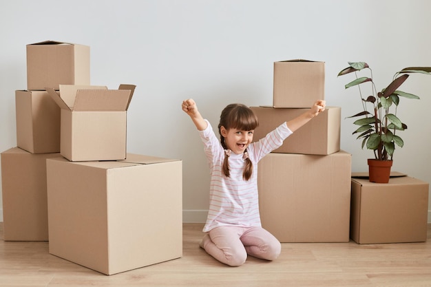 Innenaufnahme eines glücklichen, positiven kleinen Mädchens, das während des Umzugs in eine neue Wohnung in der Nähe von Kartons mit persönlichen Stapeln auf dem Boden sitzt, hob die Arme und feierte den Umzug