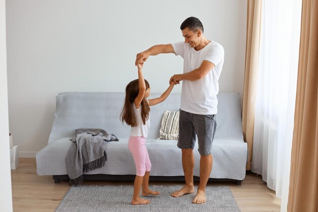 Innenaufnahme eines glücklichen Vaters, der ein kurzes und weißes T-Shirt in Jeans trägt, der mit seiner Tochter gegen das Sofa im Wohnzimmer tanzt und die Zeit mit Kind genießt.