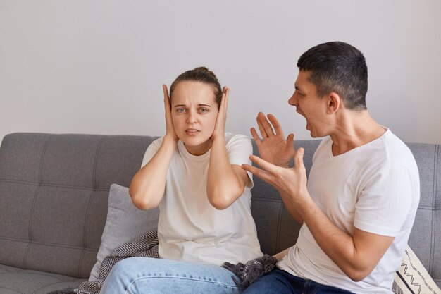 Innenaufnahme eines jungen Paares, das einen Konflikt hat, ein wütender Mann, der auf dem Sofa sitzt und seine Frau mit erhobenen Armen anschreit, die Streitprobleme in der Beziehung hat