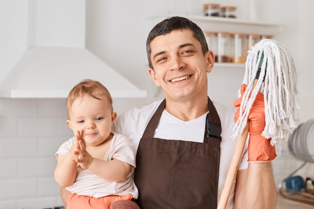 Innenaufnahme eines kaukasischen jungen erwachsenen brünetten Mannes mit Säuglingsbaby und Bodenwischern zum Reinigen der Küche mit brauner Schürze und weißem T-Shirt, der mit glücklichem Gesichtsausdruck in die Kamera schaut