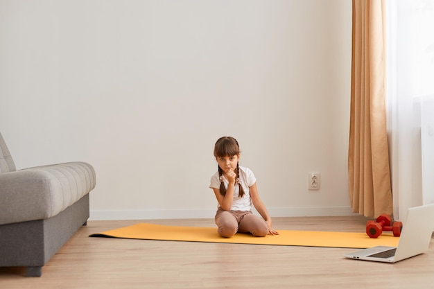 Innenaufnahme eines kleinen Mädchens, das auf einer Matte auf dem Boden sitzt und in die Kamera schaut, während es sich ausruht, während es allein zu Hause Sportübungen macht Kindergesundheit und Fitness