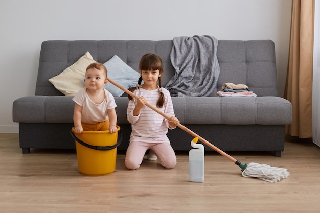 Innenaufnahme eines kleinen Mädchens mit Wischmopp in den Händen und ihrer kleinen Schwester, die gemeinsam im Wohnzimmer Haushalt macht und Spaß hat, während sie das Baby in der Wohnung aufräumt, das im Eimer sitzt