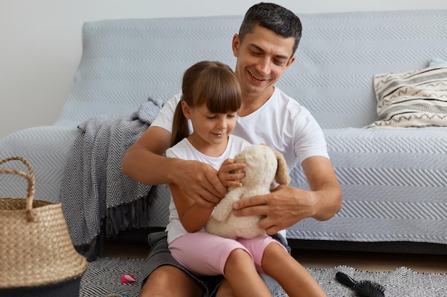 Innenaufnahme eines lächelnden attraktiven Mannes mit weißem T-Shirt im lässigen Stil, der auf dem Boden in der Nähe des Sofas sitzt und seine Tochter hält und zusammen mit einem weichen Hundespielzeug spielt.