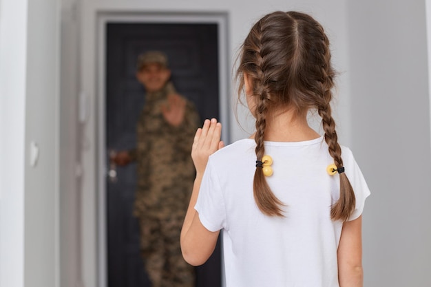 Innenaufnahme eines Militärvaters, der in den Krieg zieht, ein kleines Mädchen mit dunklen Haaren und Zöpfen, das rückwärts posiert und mit der Hand winkt, um sich vom Vater zu verabschieden, der an der Tür posiert