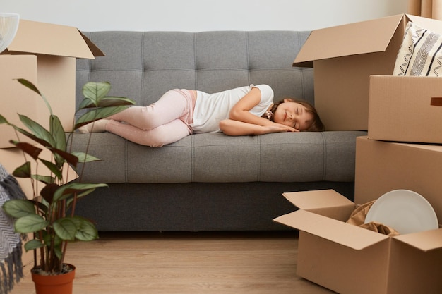 Innenaufnahme eines müden weiblichen Kindes in Freizeitkleidung, das auf dem Sofa liegt und schläft, müde und erschöpft ist, nachdem es in eine neue Wohnung gezogen ist, umgeben von Paketen und Kisten.