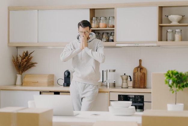 Innenaufnahme eines nachdenklichen Mannes, der am Familienbudget arbeitet, schaut aufmerksam auf Laptop-Computer-Posen in der Küche mit ausgepackten Kartons, mietet neues Haus in lässigem Sweatshirt, arbeitet aus der Ferne zu Hause