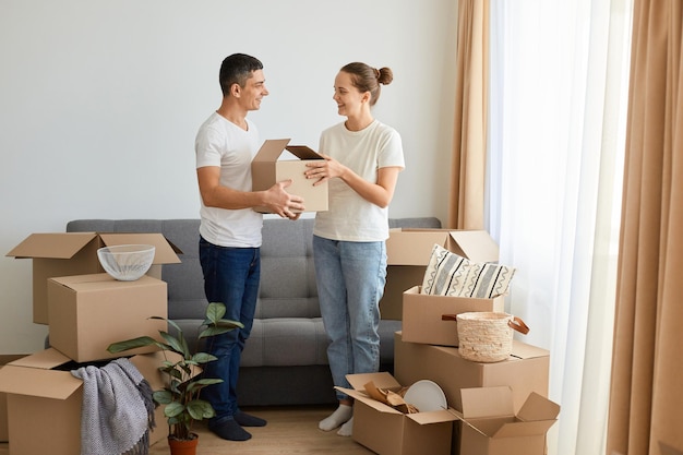 Innenaufnahme eines positiven, angenehmen jungen Paares, das während des Umzugs in der Nähe eines Sofas steht, umgeben von einer Familie aus Kartons, die positive Emotionen und Glück ausdrückt