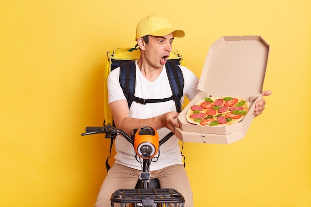 Innenaufnahme eines schockierten, erstaunten Kuriermanns auf dem Fahrrad öffnet eine Pizzaschachtel und stellt fest, dass er Bestellungen verwechselt, hält den Mund offen und trägt ein weißes T-Shirt und eine Kappe, die isoliert über gelbem Hintergrund liegen