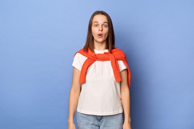 Innenaufnahme eines überraschten kaukasischen Mädchens, das ein weißes T-Shirt und einen orangefarbenen Pullover trägt, der über die Schultern gebunden ist und an einer blauen Wand steht, die mit großen Augen in die Kamera blickt, sieht etwas Erstaunen