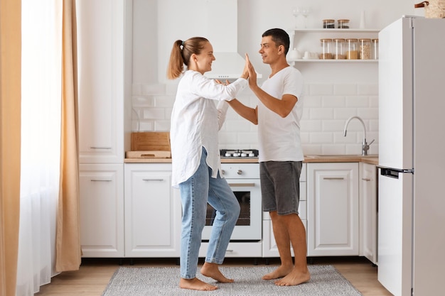 Innenaufnahme in voller Länge von positiven Paaren, jungen erwachsenen Männern und Frauen, die weiße Hemden und Jeans tragen und zu Hause gegen leichtes Küchenset zusammen tanzen.