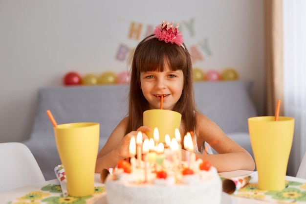 Innenaufnahme von dunkelhaarigen, lächelnden kleinen, die mit kuchen und kerzen am tisch sitzen, limonade aus gelben gläsern trinken, glückliche positive emotionen ausdrücken und in die kamera schauen.