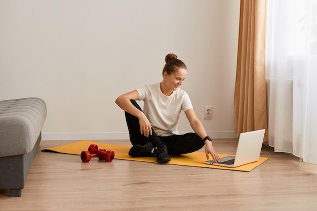 Innenaufnahme von jungen erwachsenen weiblichen Sitzübungen auf einer Yogamatte vor dem Notebook zu Hause, die Fitnessvideos auf ihrem Computer auswählt, der auf einer Yogamatte sitzt