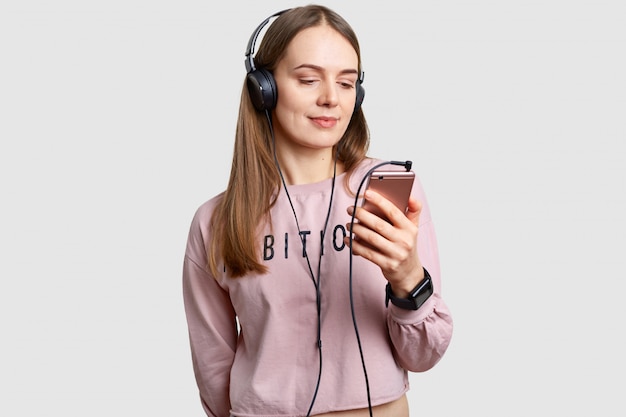 Innenaufnahme von Teenager-Mädchen im Bildschirm des Smartphones konzentriert, hört Musik mit Kopfhörern von der Wiedergabeliste, genießt lauten reinen Klang, hat lange Haare, isoliert über weiße Wand. Freizeitkonzept