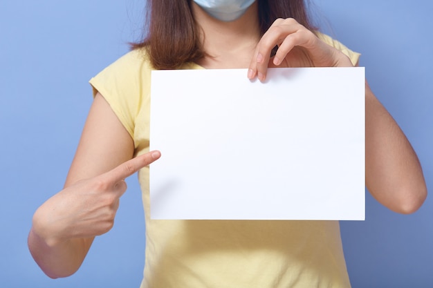 Innenbild einer unbekannten jungen Frau, die eine antibakterielle Maske trägt und ein leeres Blatt Papier hält