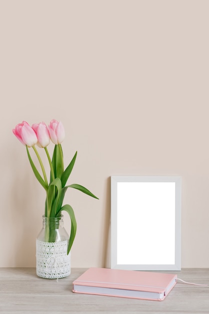 Inneneinrichtung mit dekorativen Elementen. Layout mit weißem Rahmen und rosa Tulpen in einer Vase und einem rosa Notizbuch auf dem Tisch auf hellbeigem Hintergrund