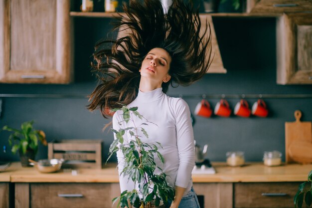 Innenlebensstilporträt des schönen jungen Mädchens mit verstreutem Haar und gekipptem Kopf, der Topf mit Blume hält.