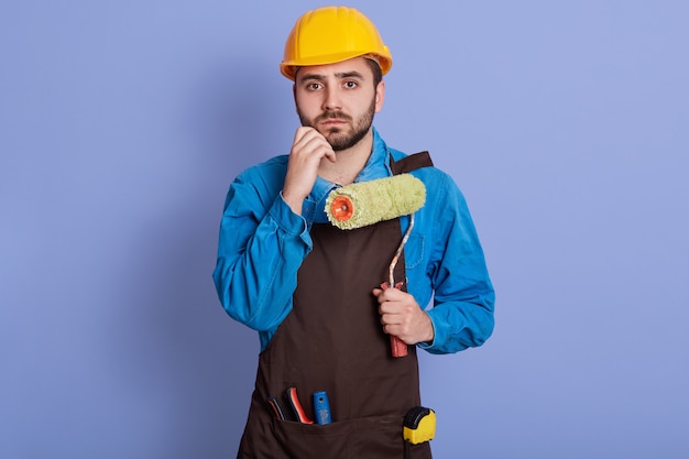 Innenporträt des enttäuschten verwirrten jungen Baumeisters, der Hand an Kinn legt, Walze hält, nachdenklichen Gesichtsausdruck hat, Uniform und Helm trägt und isoliert über lila Wand posiert.