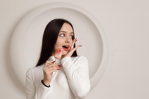 Innenporträt einer positiv lächelnden, brünetten jungen Frau auf weißem Hintergrund, die süßen Dessertkuchen in den Händen hält