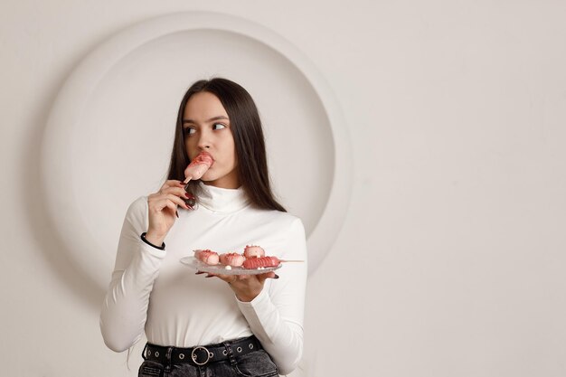 Innenporträt einer positiv lächelnden, brünetten jungen Frau auf weißem Hintergrund, die süßen Dessertkuchen in den Händen hält