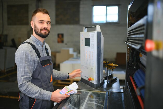 Innenraum des Druckhauses mit moderner Offsetdruckmaschine und Bediener in Arbeitsuniform, der die Qualität des Drucks überprüft und den Druckprozess kontrolliert