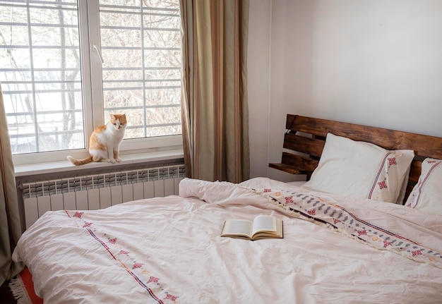 Innenraum des Schlafzimmers Leichtes Bettzeug Buch auf dem Bett und Katze am Fenster