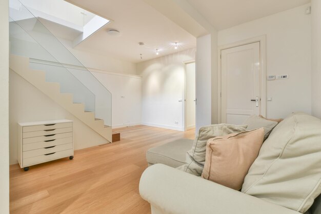Innenraum einer leeren, hellen Halle mit weißem Sofa und Kissen darauf, Schrank und einer Treppe zum Obergeschoss