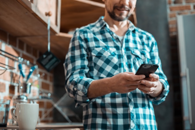 Innovador smartphone en manos de un buen hombre guapo positivo mientras está de pie en la cocina