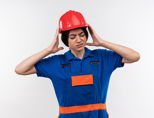 Insatisfecho con los ojos cerrados joven constructor mujer en uniforme tomados de la mano alrededor de la cabeza