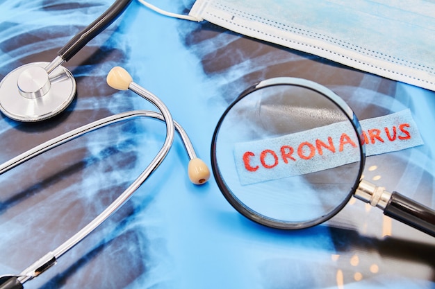 Inschrift Coronavirus durch eine Lupe auf Hintergrund von Stethoskop, medizinischer Maske und Lungenröntgen. Nahansicht