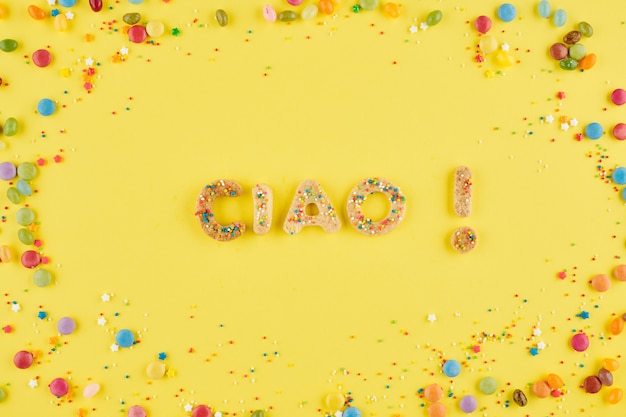 Inscripción Ciao hecha de dulces galletas caseras sobre fondo amarillo con caramelos de chocolate y chispitas de colores