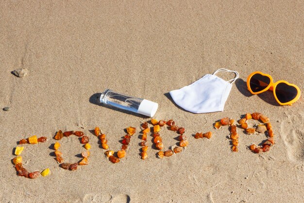 Foto inscripción covid19 hecha de piedras de ámbar máscara protectora líquido desinfectante y gafas de sol en la arena en la playa vacaciones seguras durante el coronavirus