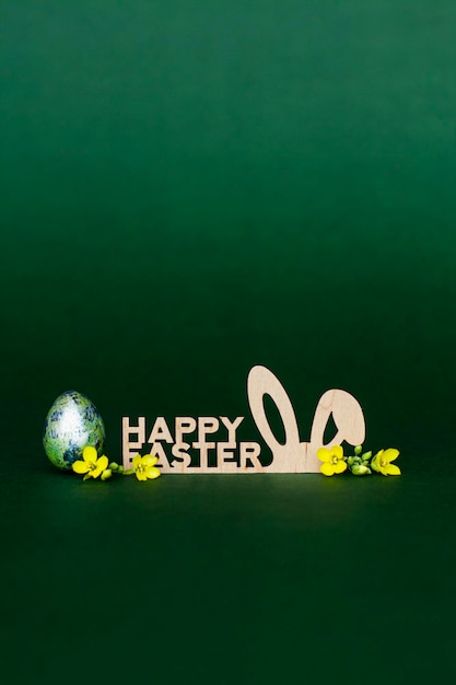 Foto inscripción de madera feliz pascua orejas de conejo estilizadas sobre un fondo verde oscuro huevo de codorniz y pequeñas flores amarillas
