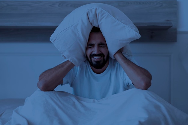 Foto insônia estressou o homem indiano cobrindo os ouvidos com travesseiro enquanto está sentado na cama