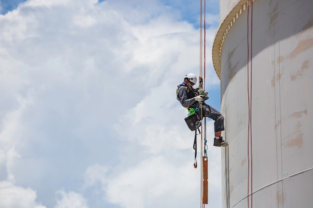 Inspeção do trabalhador masculino usando a primeira linha de segurança da corda do arnês de segurança trabalhando em um lugar alto no céu azul de gás esférico do teto do tanque