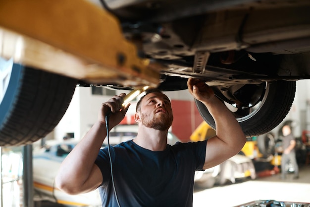 Inspección de la antorcha y mecánico con el vehículo en el taller para el mantenimiento de servicio o reparación para la seguridad Ingeniería automotriz ligera y trabajador industrial masculino comprobando el fondo del coche para detectar fugas de aceite en el garaje