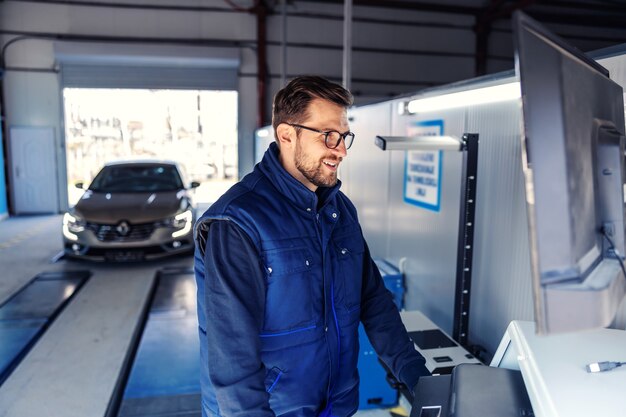 Inspección técnica del coche. Diagnóstico de automóviles en inspección técnica, electrónica de automóviles. Un hombre con uniforme azul se para frente a la computadora y ajusta los parámetros de visualización de la pantalla