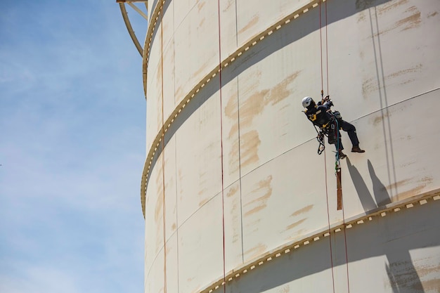 Inspección del trabajador masculino usando la línea de seguridad de la cuerda del arnés de seguridad primero trabajando en un lugar alto en el cielo azul de gas esférico del techo del tanque