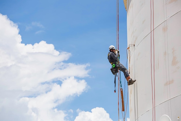 Inspección del trabajador masculino usando la línea de seguridad de la cuerda del arnés de seguridad primero trabajando en un lugar alto en el cielo azul de gas esférico del techo del tanque