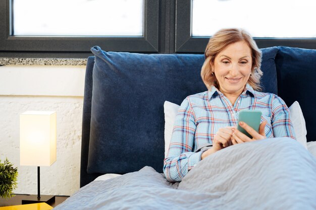 Inspecionando smartphone. Mulher adulta concentrada encostada na cama e verificando as redes sociais durante a rotina matinal normal