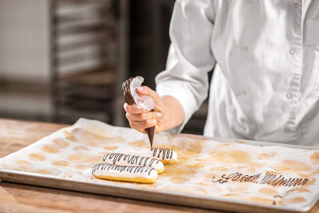 Inspiração, chef pasteleiro. Confeiteiros espremendo um saco de confeitar com chocolate sobre um eclair fresco deitado na assadeira